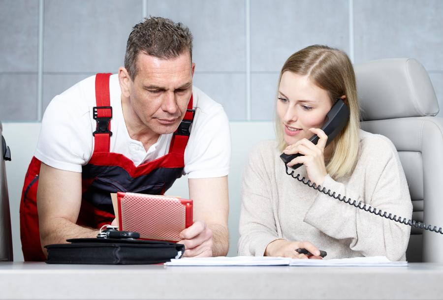 Frau am Telefon, neben ihr steht ein Mechaniker mit Formularen