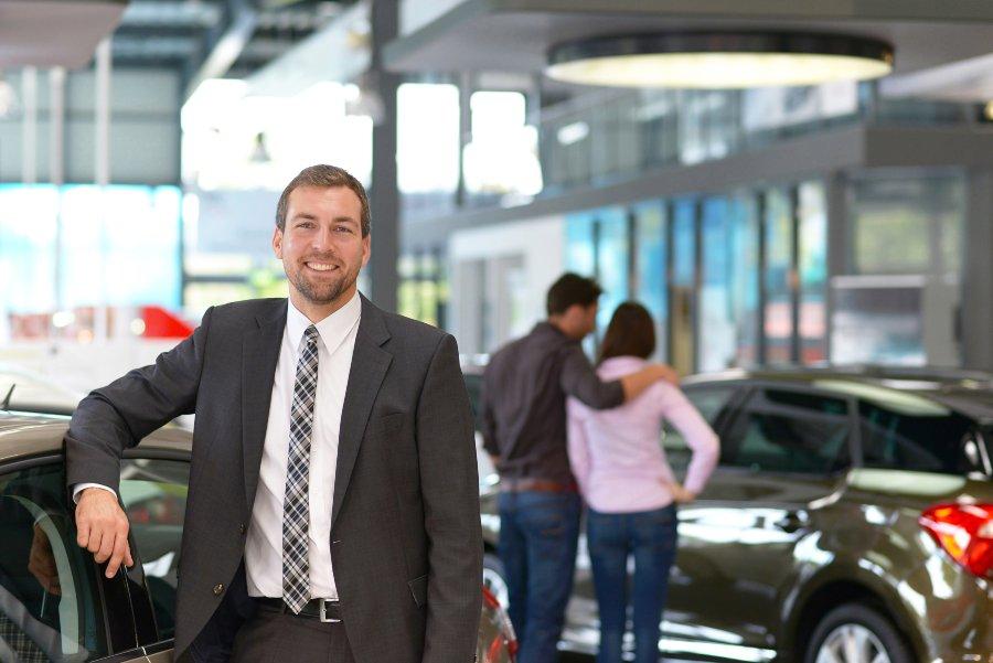 Ein Autoverkäufer lächelt im Autohaus in die Kamera, hinter Ihm kauf ein junges Paar ein Neuwagen Kundengewinnung