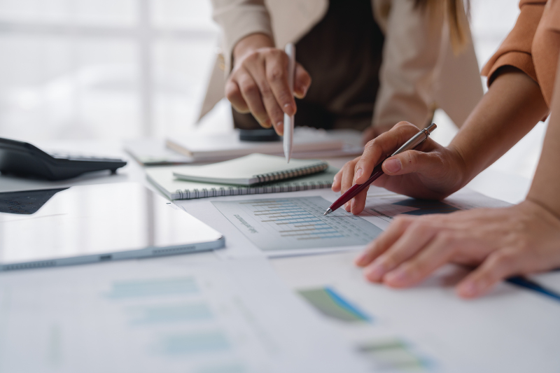 Blick auf einen Schreibtisch, auf dem viele Papiere und Kalender ausgebreitet sind. Zwei Personen deuten mit Kugelschreibern in den Händen auf einzelnen Papiere. 